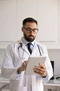 Laboratuvar önlüklü, şık gözlüklü, tıbbi işler için dijital cihaz kullanan, hastalarla sohbet eden, elektronik kayıtları okuyan ciddi meşgul İspanyol doktor.