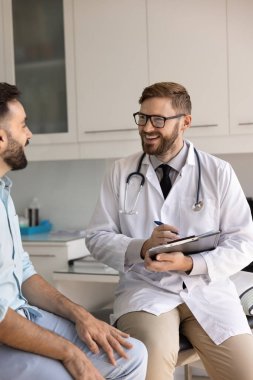 Pozitif genç bir doktor hastasına sağlık sigortası, hastalık belirtileri, sağIık durumu, not defterine not yazma, anketi doldurma, gülümseme, gülme, tıbbi kanepede oturma gibi konuları soruyor. Dikey çekim