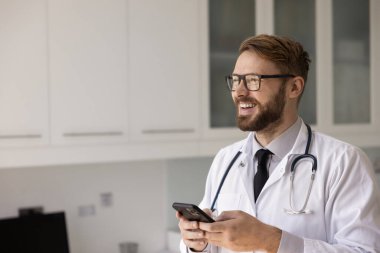 Gözlüklü ve laboratuvar önlüklü genç bir pratisyen. Çalışma odasında akıllı telefondan fotokopi odasına bakıyor, gülümsüyor, gülüyor. Doktor cep telefonunda hastalarla sohbet ediyor.