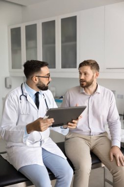 Ciddi Latin pratisyen doktor erkek hastaya sağlık şikayetleri, dinleme, tıbbi kayıtları, geçmişi olan defteri tutma, klinik muayene odasında danışmanlık yapma