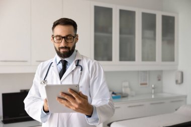 Gözlüklü ve steteskoplu genç Latin pratisyen doktor tablet üzerindeki elektronik tıbbi raporları karıştırıyor, hastalarla çevrimiçi iletişim kurmak için dijital cihaz kullanıyor.