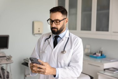 Ciddi meşgul erkek doktor muayene odasında cep telefonuyla yazı yazıyor, hastalarla sohbet ediyor, çevrimiçi destek veriyor, yardım ediyor, sağlık hizmeti veriyor, iletişim için akıllı telefonu kullanıyor.