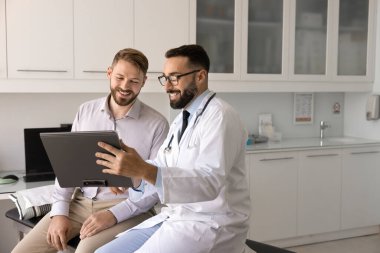 İyimser bir erkek pratisyen hastaya tıbbi muayene sonuçlarını açıklıyor. Kağıt üzerinde kağıt dökümanlar gösteriyor, gülümsüyor, muayene için klinik kanepesinde oturuyor, danışmanlık yapıyor.