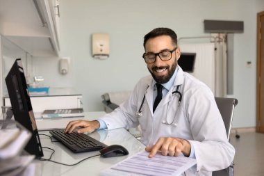 Laboratuvar önlüklü ve gözlüklü pozitif erkek pratisyen klinik ofisindeki çalışma masasında tıbbi kayıtları okuyor, dosyalara, belgelere, kağıt incelemelerine iyi sonuç veriyor, gülümsüyor.