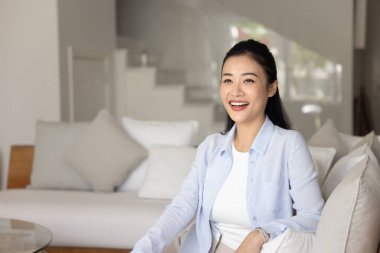 Keyifli bir kadın, rahat ve modern mobilyaları olan şık oturma odasındaki koltukta oturmuş gülüyor, mutlu bir gülümsemeyle uzaktan bakıyor, hafta sonu evde mutluluk ve haz yayıyor.