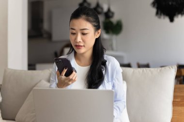 Asyalı bir kadın akıllı telefonunu kullanarak kanepeye oturur, sosyal medyaya göz atar, e-postalarını kontrol eder, serbest çalışmasıyla ilgili haberleri ya da makaleleri okur, yeni cep telefonu uygulaması kullanır ya da internetten bilgi toplar.