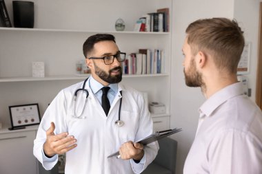 Kendine güvenen erkek doktor hastaya tıbbi tavsiyeler veriyor, hastane muayene odasında genç bir adamla konuşuyor, teşhisi ve tedaviyi açıklıyor.