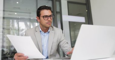 Genç profesyonel Latin adam dizüstü bilgisayarını kullanarak ve belgelerden veya materyallerden elde edilen verileri analiz ederek, basılı araştırma ve notlara dayalı sunum hazırlayıp, sözleşmeleri veya yasal belgeleri doğrulamak