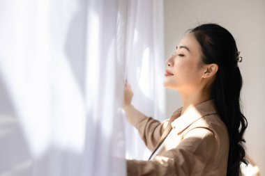 Huzurlu Asyalı bir kadının pencerenin yanında durup, beyaz perdeleri açması, temiz havanın tadını çıkarması gözleri kapalı ve derin derin düşüncelere daldığı huzurlu bir uyanış anı. Yeni güne hoş geldin.