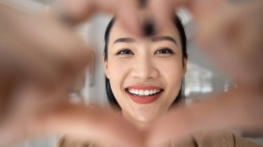 Kameraya kalp şeklinde el hareketlerini gösteren gülümseyen Asyalı kadının geniş bir görüntüsünü kapatın. Sevgisini ve sevgisini ifade ediyor. Diş bakımı, ortodontoloji ve sigorta hizmetlerinin reklamını yapıyor.