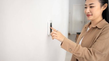 Beyaz duvarın yanında duran kadın, duvara monte edilmiş dijital termostat veya akıllı kontrol panelinde düğmeye basıyor, sıcaklığı ayarlıyor, ev otomasyonunu yönetiyor, ısıtmayı, soğutmayı, güvenlik sistemini ayarlıyor