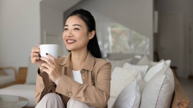 Serene Vietnamlı kadın dinleniyor, bir fincan taze çay ya da kahveyle kanepede dinleniyor, gülümsüyor, evde kaygısız anların tadını çıkarıyor en sevdiği, lezzetli içeceklerle oturma odasında.
