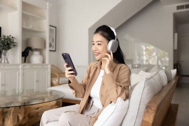 Rahat bir kanepede dinlenen, kulaklıklarıyla müzik veya podcast dinleyen, akıllı telefondan video izleyen, internette boş zaman geçiren, video çağrısına katılan, dijital iletişimde yer alan bir kadın.