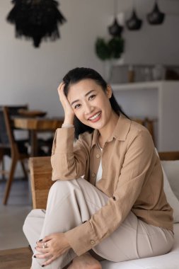 Modern oturma odasındaki rahat koltukta rahat rahat dinlenen rahat giyinmiş gülümseyen Asyalı kadın, kameraya bak. Tatmin olmuş ev sahibi ya da kiracısının portresi boş zamanlarını kendi evinde ya da kiralık evde geçiriyor