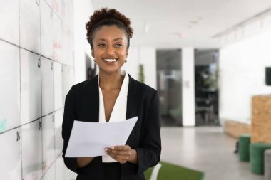 25 'li yaşlardaki Afrikalı genç iş kadını, proje yöneticisi ya da idari işçi portresi, plan, strateji, yapılacaklar listesi, içgörü ya da programla birlikte beyaz duvar tahtasının yanındaki modern çalışma ofisinde poz veriyor.