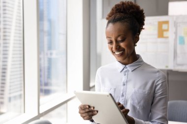 Modern tablet cihazıyla çalışan gülümseyen Afrikalı iş kadını, iş sunumu veya pazarlama stratejisi hazırlıyor, programları yönetiyor veya takvim uygulaması aracılığıyla toplantılar düzenliyor
