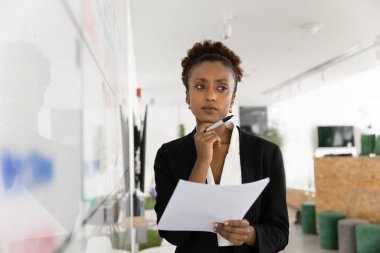 Ciddi Afrikalı profesyonel bir kadın kağıda yazılmış bilgileri değerlendiriyor, karar vermek için bilgileri gözden geçiriyor, meydan okuma yoluyla düşünüyor, planlama stratejisi kuruyor, büyük beyaz tahtanın yanında duruyor.