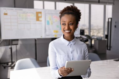 Gülümseyen Afrikalı kadın, modern toplantı odasında elinde tabletle duran ofis çalışanı, mutlu bir gülümsemeyle uzaklığa bakıyor, çevrimiçi çalışıyor, proje yönetim araçlarını veya görev listelerini uygulama kullanarak güncelliyor