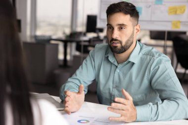 Profesyonel adam masadaki çizelgelerde sunulan bilgileri, iş stratejisi veya proje planının bir sonraki adımlarını özetleyerek, fikir alışverişinde bulunarak, ofiste toplanan müşteri veya meslektaşlarına bakış açısını anlatıyor.