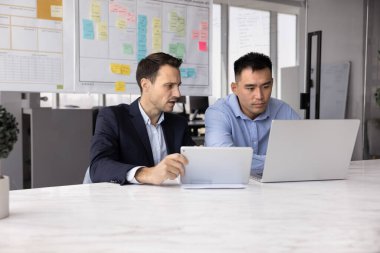 Asyalı meslektaşı dizüstü bilgisayarda çalışırken İspanyol adam dijital tablet kullanıyor, ortak görevlerine odaklanmış vaziyette, ofiste iki modern cihaz kullanarak işbirliğine giriştiler. Dijital iş akışı