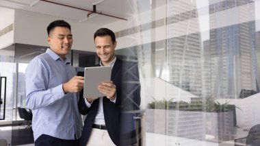 İki gülümseyen erkek ekip üyesi, birlikte tablet kullanarak, düzenleyerek veya belgeyi gözden geçirerek, web sitesi veya çevrimiçi kaynak üzerinden bilgi arayarak, proje yönetim yazılımına erişerek,