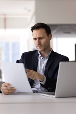 Odaklanmış iş adamı, sözleşmenin öğrenme noktalarını dikkatle okuyor, modern işyerindeki açık dizüstü bilgisayarla masada oturan resmi anlaşmada belirtilen önemli ayrıntıları ve sorumlulukları analiz ediyor.