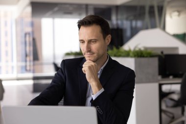 40 'lı yaşlarda, ceketli İspanyol profesyonel bir adam masada oturmuş laptopa ciddi, düşünceli bir ifadeyle bakıyor, işle ilgili haberleri inceliyor, belgelerini okuyor ya da proje güncellemelerini okuyor.