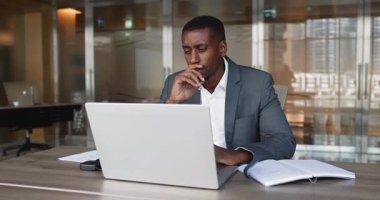 Düşünceli Afrikalı Amerikalı şirket yöneticisi bilgisayarda çalışıyor, ofiste büyük bir masada oturuyor, çeneye dokunuyor, başka tarafa bakıyor, alete sarılıyor, dizüstü bilgisayarında daktilo yazıyor.