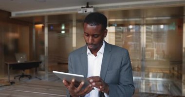 Ciddi odaklı Afrikalı iş adamı resmi takım elbise içinde finansal banka hizmetlerini tablet üzerinde kullanıyor, dijital cihaz üzerinde yazıyor, internette geziniyor, online ürün üzerinde çalışıyor, ofiste duruyor.