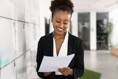 Afrikalı iş kadını elinde çarşaf tutuyor, kağıt üzerinde yapılacaklar listesini gözden geçiriyor, randevularını ya da son teslim tarihlerini kontrol ediyor, bilgileri karşılaştırıyor, gülümsüyor, organize programdan memnun.