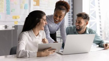 Üç neşeli iş arkadaşı modern ofiste toplanıyor, online görevi gözden geçiriyorlar, birlikte çalışıyorlar, birlikte gülüyorlar, yaratıcı çözümleri paylaşıyorlar, başlangıç veya proje güncellemeleri için fikirler, iş yerinde takım çalışması yapıyorlar.