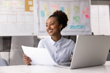 Afrikalı kadın çalışan, elinde dizüstü bilgisayarından çıkarılmış belgelerle masada oturuyor, mutlu bir gülümsemeyle mesafeye bakıyor, olumlu geri bildirim ya da sonuçları değerlendiriyor, mesleki gelişimden tatmin oluyor.