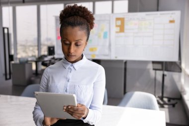 Ciddi Afrikalı iş kadını, konferans odasında tek başına duran tablet kullanıyor, müşteri e-postalarına cevap veriyor, işle ilgili konuları uzaktan çözüyor, sözleşmeleri veya teklifleri gözden geçiriyor, bilgi araştırıyor.