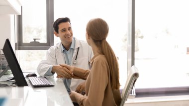 Genç ve mutlu bir Latin doktor kadın hastayla tokalaşıyor, iyileşme için tebrik ediyor, test sonuçları iyi, klinikteki işyerinde oturuyor, gülümsüyor, konsültasyonu bitiriyor. Sancak vuruşu