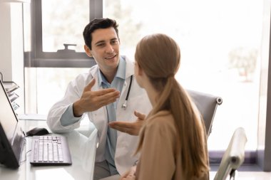 Pozitif genç İspanyol sağlık uzmanı klinikteki danışma toplantısında kadın hastaya tıbbi danışmanlık yapıyor, işyerinde oturuyor, kadınlarla konuşuyor, gülümsüyor.