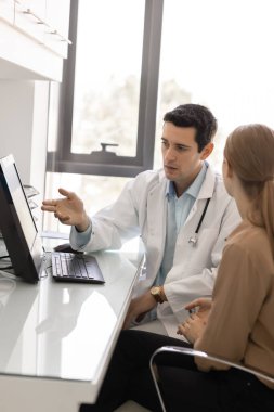 Laboratuvar önlüklü, iş yerindeki kadınla konuşan, bilgisayar monitöründeki elektronik sağlık raporuna işaret eden, teşhisi açıklayan, muayene sonuçlarını kadın hastaya açıklayan ciddi bir İspanyol doktor. Dikey çekim