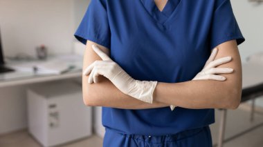 Mavi önlüklü ve lastik eldivenli genç bir cerrah vuruldu. Kadın sağlık uzmanı, doktor, hemşire, cerrah kadın klinik muayene odasında elleri katlanmış bir şekilde duruyor.