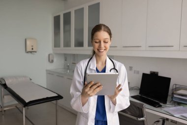 Neşeli genç doktor, hastalarla tele-sağlık hizmetlerinde sohbet ediyor, klinikte tablet yazıyor, online destek veriyor, danışmanlık yapıyor, muayene odasında dijital cihazlar kullanıyor.