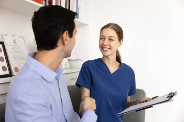 Mavi önlüklü genç bayan doktor erkek hastayla el sıkışıyor, elinde dokümanlar, klinik başvuru formu, tıbbi testler iyi sonuçlar, gülümsüyor, adama el sıkışıyor.