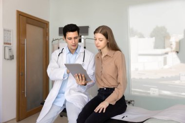 Ciddi genç İspanyol doktor, sağlık raporunu, teşhisi, elektronik muayene sonuçlarını, hastane muayene odasında bir hastayla konuşurken anlatıyor.