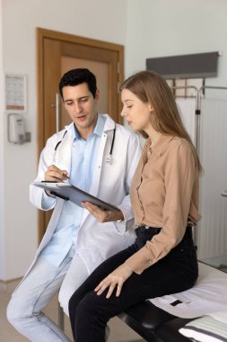 Kadın hastaya reçeteyi açıklayan ciddi bir tıp uzmanı, panodaki kağıda işaret eden, konuşan, klinik kanepesinde oturan, tedavi için tavsiye veren. Dikey çekim