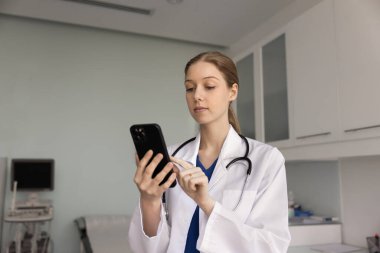 Hastane muayene odasında cep telefonuyla yazı yazan, hastalara online danışmanlık yapan, uzaktan tıbbi destek için tele sağlık uygulaması kullanan odaklanmış genç doktor kadın.