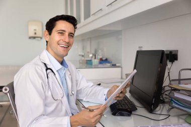 Neşeli genç İspanyol doktor hasta kayıtlarını inceliyor, elinde tıbbi raporlarla kalem ve klasör tutuyor, klinik belgelerini tutuyor, bilgisayarla iş yerinde oturuyor, dişlek gülümsemeyle başka tarafa bakıyor.