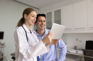Neşeli kadın pratisyen ve erkek hasta tabletten medya içeriğini izliyorlar, elektronik sağlık raporlarını tartışıyorlar, iyi kontrol sonuçları. Doktor konsültasyon için dokunmatik ped kullanıyor.