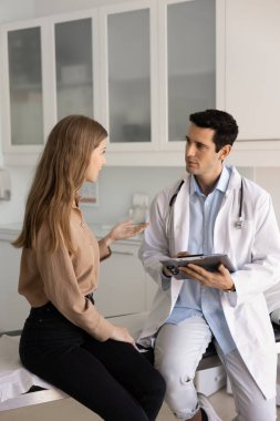 Laboratuvar önlüklü genç bir doktor hasta şikayetlerini dinliyor elinde tıbbi geçmişi, kayıtları olan bir pano tutuyor muayene odasında kanepede oturuyor ve kadınlarla konuşuyor. Dikey çekim