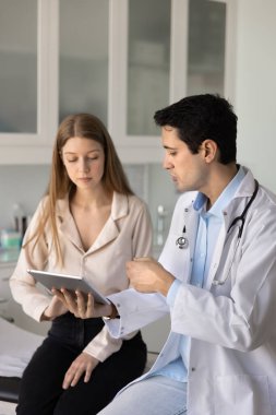 Ciddi bir Latin pratisyen doktor genç bayan hastaya tablet üzerinde elektronik kontrol sonucu gösteriyor. Hastane kanepesinde oturan kadınlara sağlık raporlarını açıklıyor. Dikey çekim