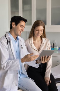 Laboratuvar önlüklü genç doktor, kadın hastayla tablet üzerinde tarama sonuçlarını tartışıyor klinik muayene odasında kanepede oturuyor, kadınlarla konuşuyor, gülümsüyor. Dikey çekim