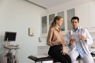 Genç, pozitif Latin pratisyen tavası tablette ultrason görüntüsünü kadın hastaya gösteriyor, tıbbi kanepede oturuyor, teşhisi açıklıyor, muayene sonuçlarını açıklıyor. Kopyalama alanı ile geniş çekim