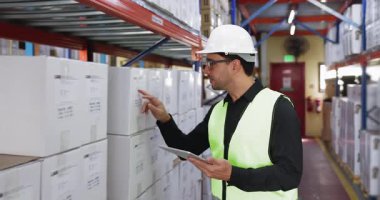 Güvenli yelek ve kask giyen, tablet kullanan, modern depolama tesisinde raflardaki etiketli kutuları inceleyen, envanter kontrolü yapan, depo operasyonları yapan Latin erkek lojistik uzmanı.
