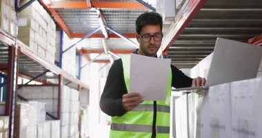 Depoda güvenlik yeleği giymiş genç Latin bir adam duruyor. Etrafı kutularla dolu raflarla çevrili. Belgeleri dizüstü bilgisayardaki verilerle karşılaştırıyor.